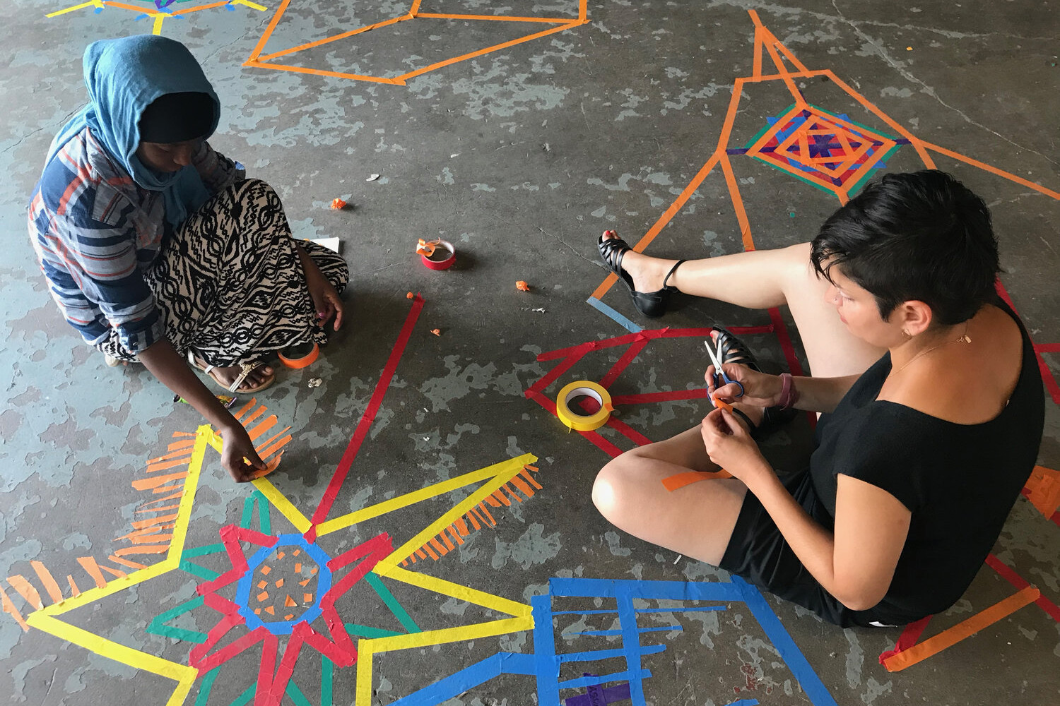 Two students sitting on the ground making designs using pieces of colored tape