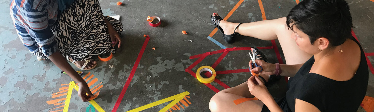 Two students sitting on the ground making designs using pieces of colored tape