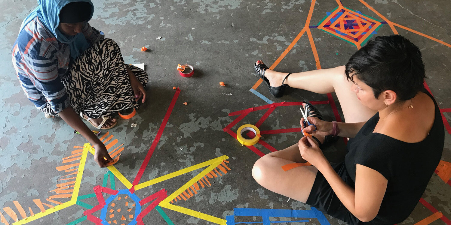 Two students sitting on the ground making designs using pieces of colored tape